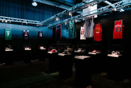 Dark exhibition showcasing hanging jerseys and shoes displayed on pedestals